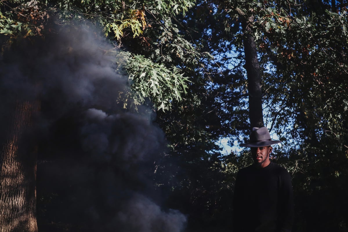 a person in a hat standing in front of trees and black smoke